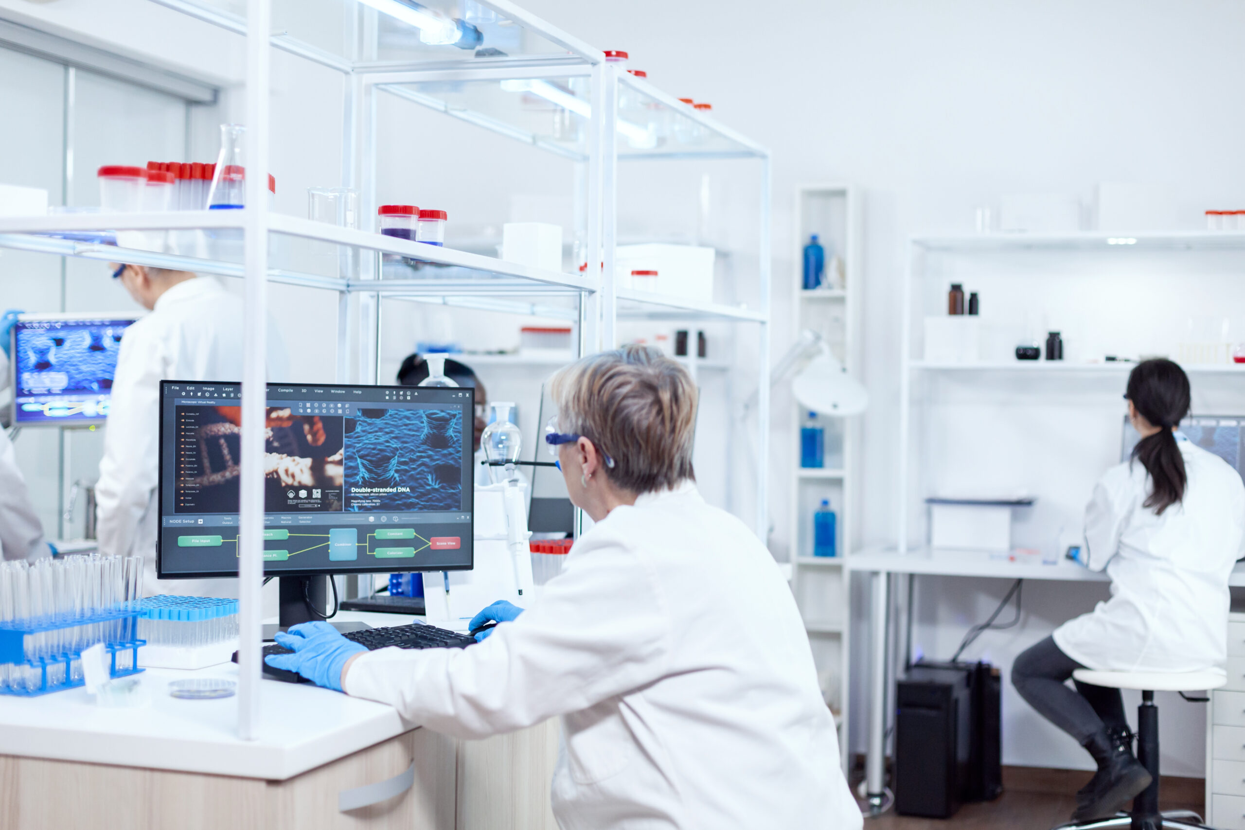 Elderly researcher working to find a cure during global pandemic Senior scientist in pharmaceuticals laboratory doing genetic research wearing lab coat with team in the background.