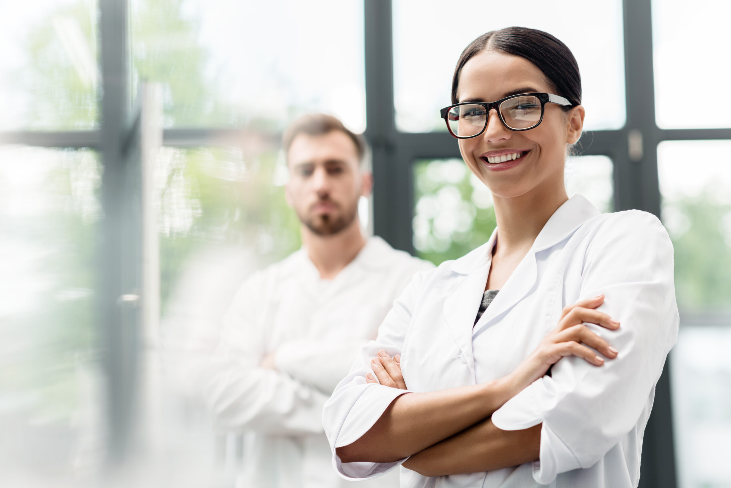 Beautiful young scientist in eyeglasses smiling at camera while colleague standing behind Beautiful young scientist in eyeglasses smiling at camera while colleague standing behind